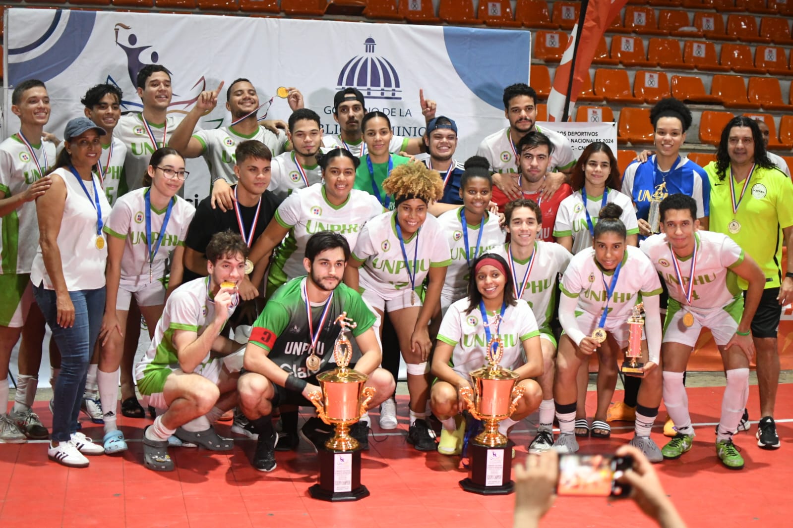 Juan Vila, viceministro de Deportes Universitarios, entrega a Aquilina Figueroa, directora de Deportes de la Universidad Nacional Pedro Henríquez Ureña (UNPHU), el trofeo de campeón del torneo de fútbol sala masculino, en la versión 2024 de los Juegos Universitarios.